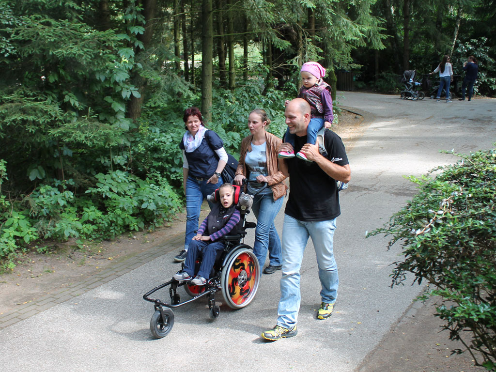 Zwei Frauen, ein Mann mit Kind auf den Schultern und ein Kind im Rollstuhl auf einem asphaltierten Weg durch einen grünen Wald