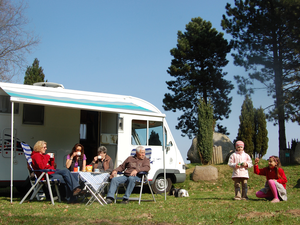 Eine Familie sitzt vor einem Wohnmobil und trinkt Café und 2 Kinder spielen auf dem Rasen