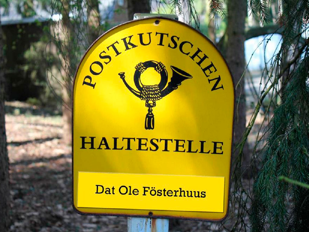 Gelbes Schild mit der Aufschrift Postkutschen Haltestelle und einem Posthorn-Symbol