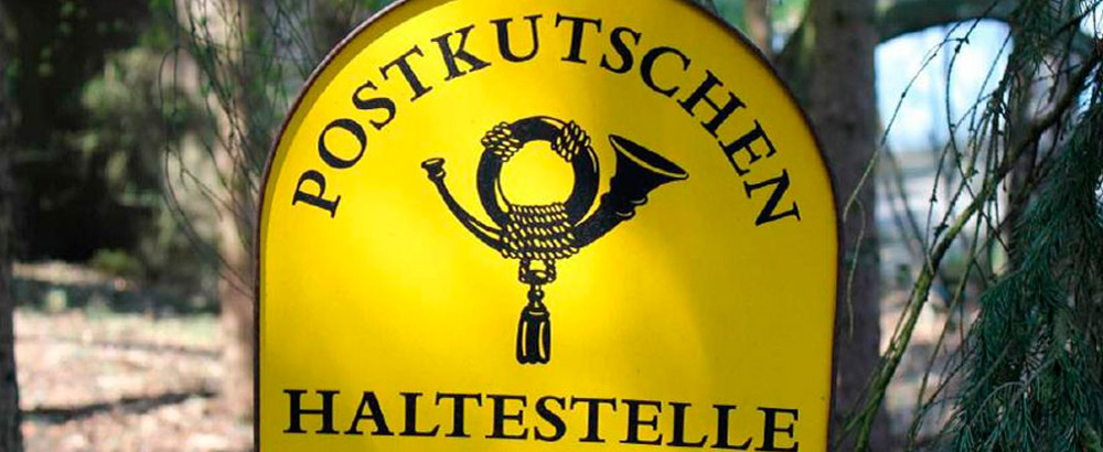 Gelbes Schild mit der Aufschrift Postkutschen Haltestelle und einem Posthorn-Symbol